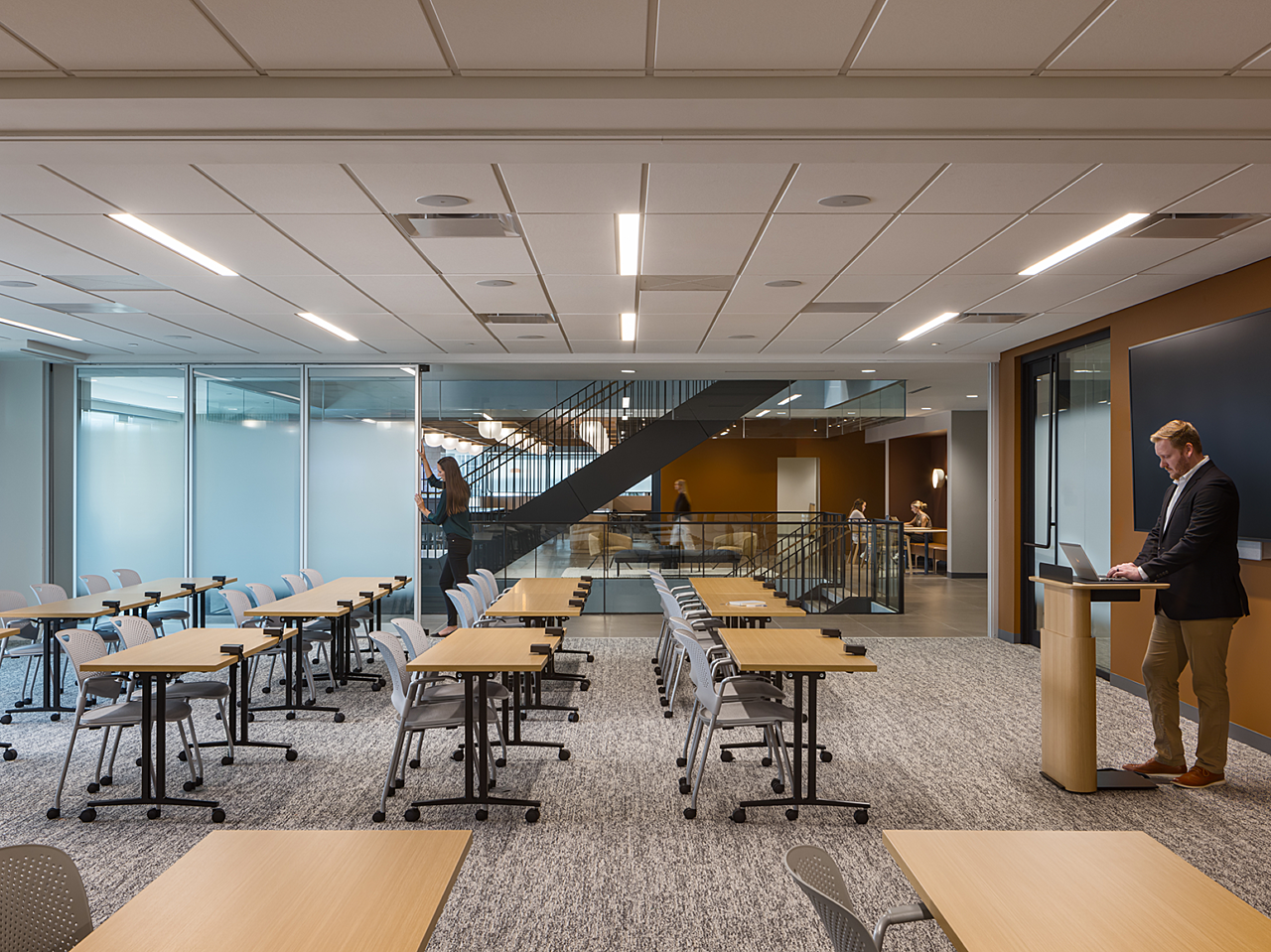 Corporate training room with moveable, frosted walls. Mobile tables and chairs. Man works on laptop at podium while woman opens the wall.