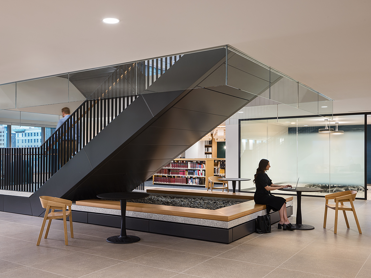 Large black staircase with its base lined with a wooden bench. Black cafe tables and wooden cafe chairs are around it. A woman works on a laptop at one.
