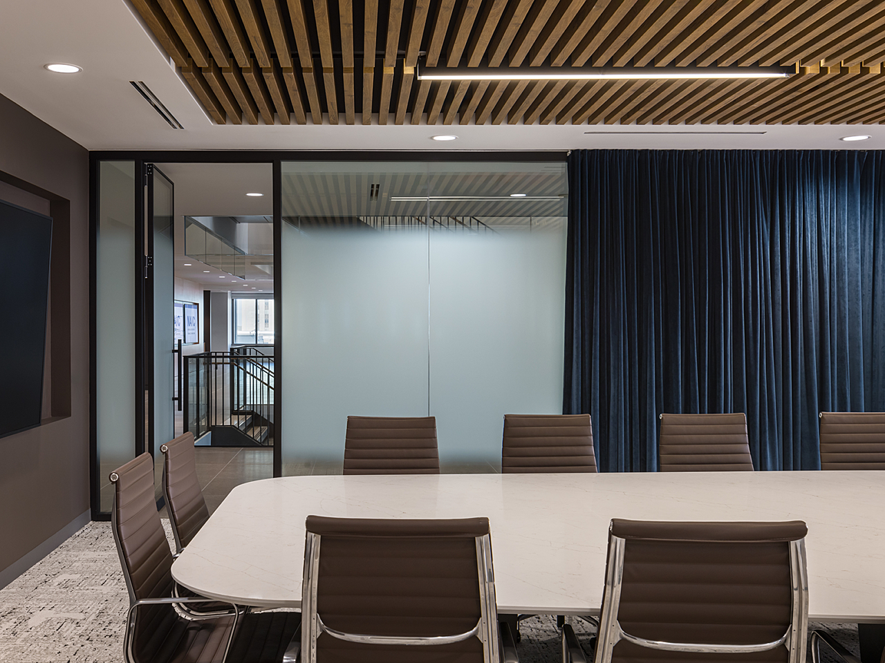 Conference room with brown, highback chairs and white table. Wooden slats line the ceiling. Frosted glass wall/door with dark blue curtains for privacy