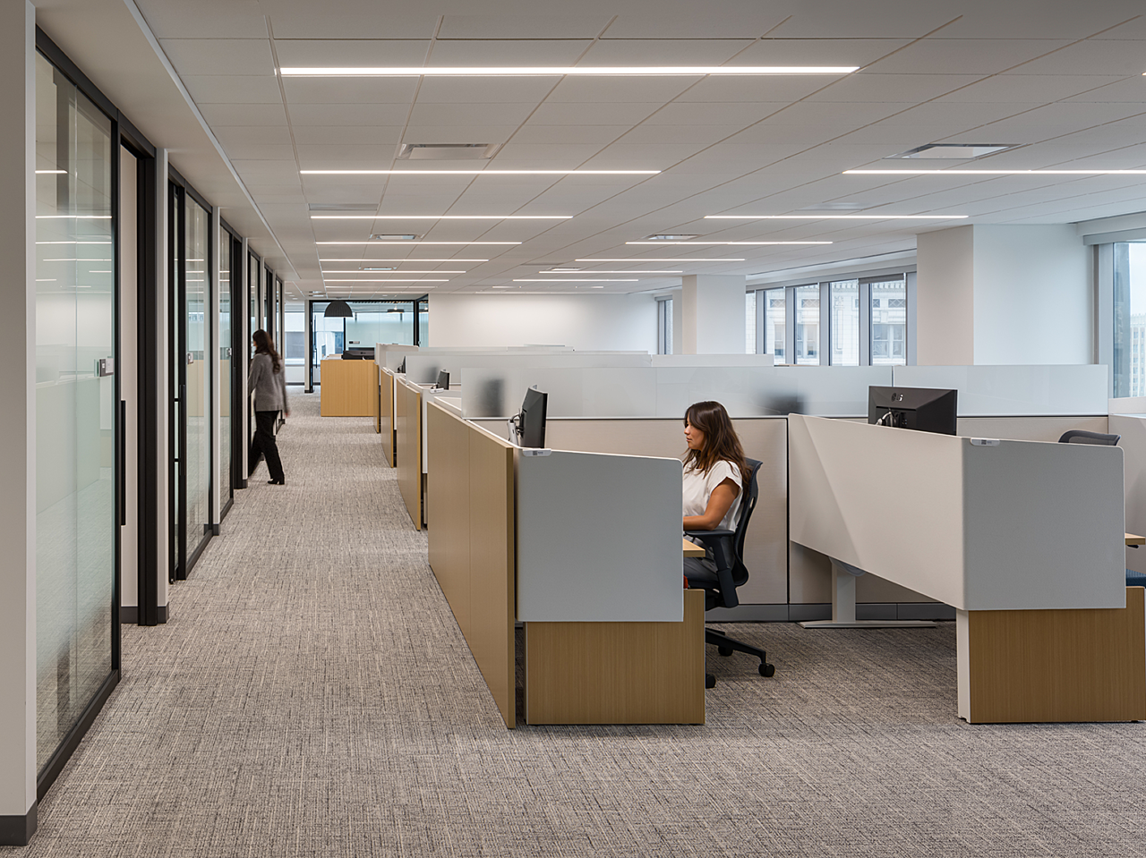 Workstations in front of a row of private, glass-fronted offices. Workstations have wood/white privacy panels that wrap to the sides. Frosted glass between