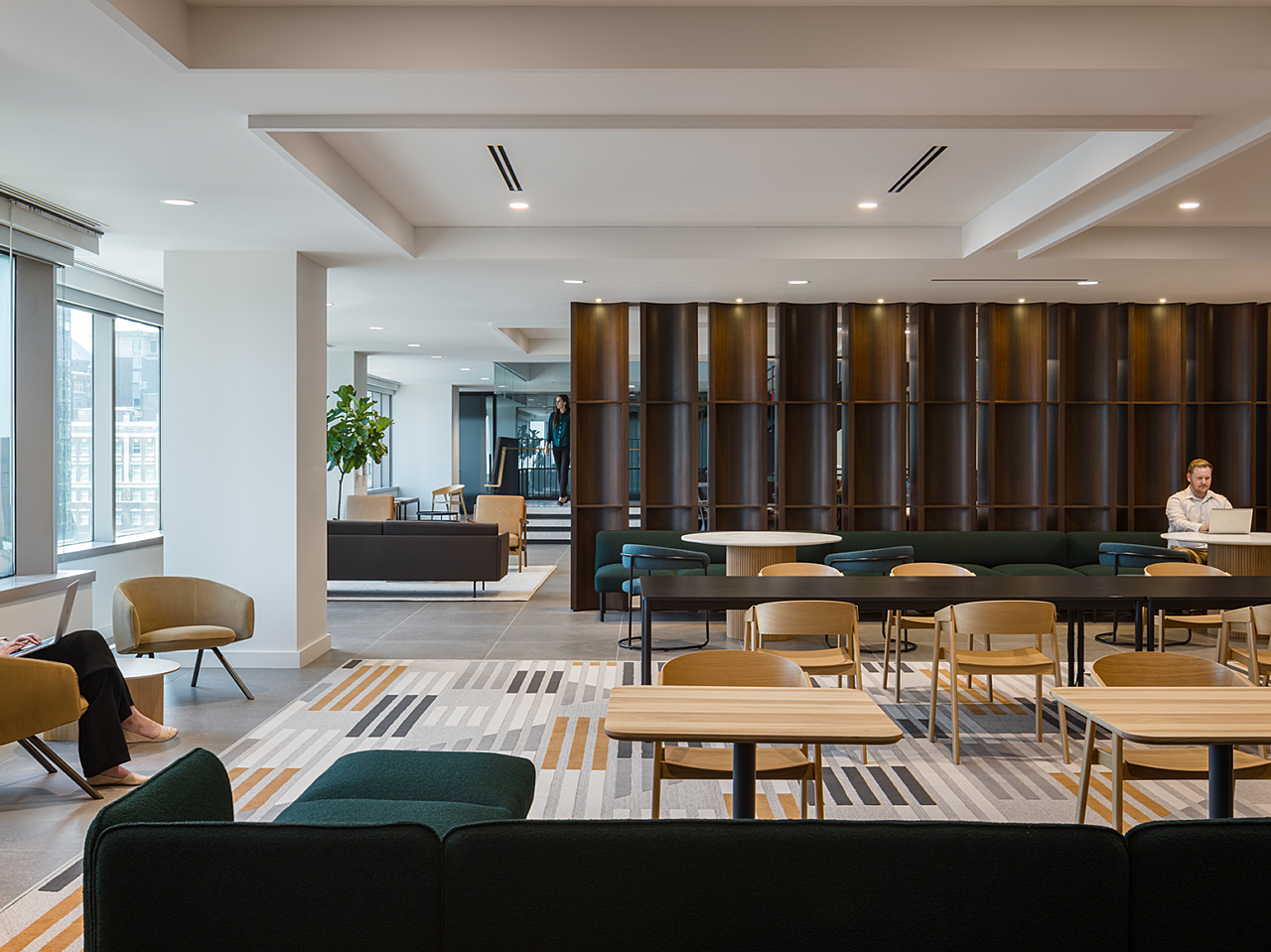Open lounge  area with large gold/black/white/grey rug, banquette seating at long and cafe tables. Large, dark wooden panel divides another open area.