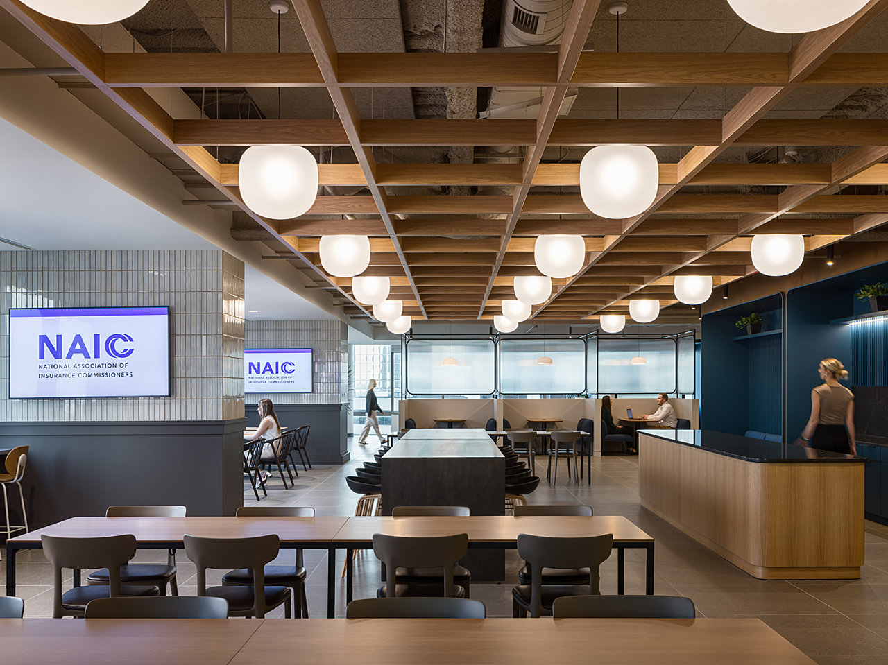 Modern breakroom with wooden tables with contrasting chairs, blue and white tile, bulb lights. Employees sit at booths and tables and others walk space