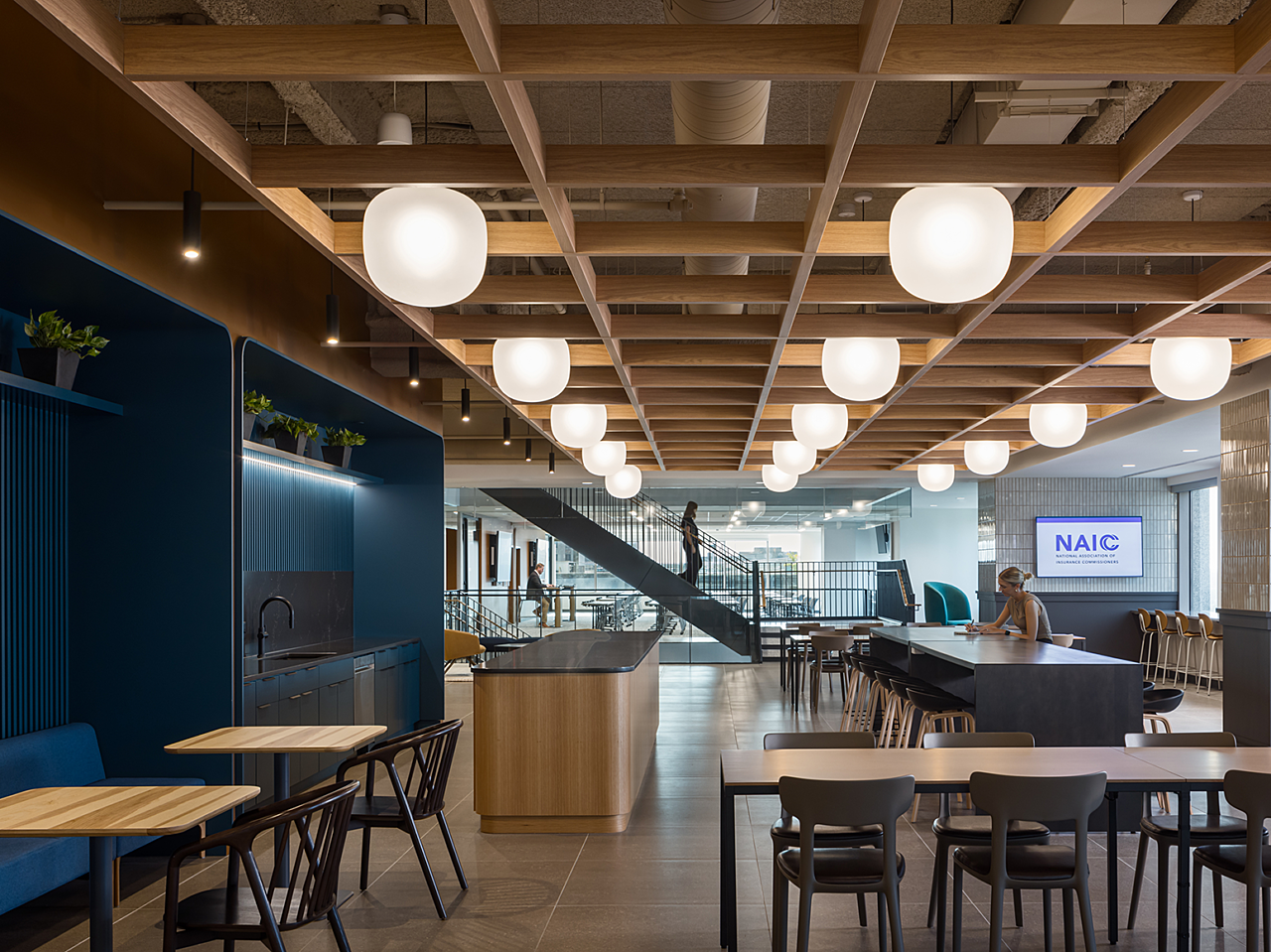 Breakroom with dark blue walls and accents, white tile, and various long, tall and cafe tables with chairs and stools. A stairwell shows in background.