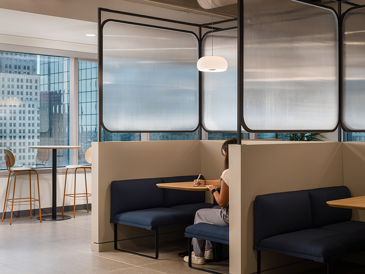 Cafe booths with 3-sided walls, blue mid-back benches and frosted partitions and a drop light. Tall cafe table near windows. Woman works in one booth.