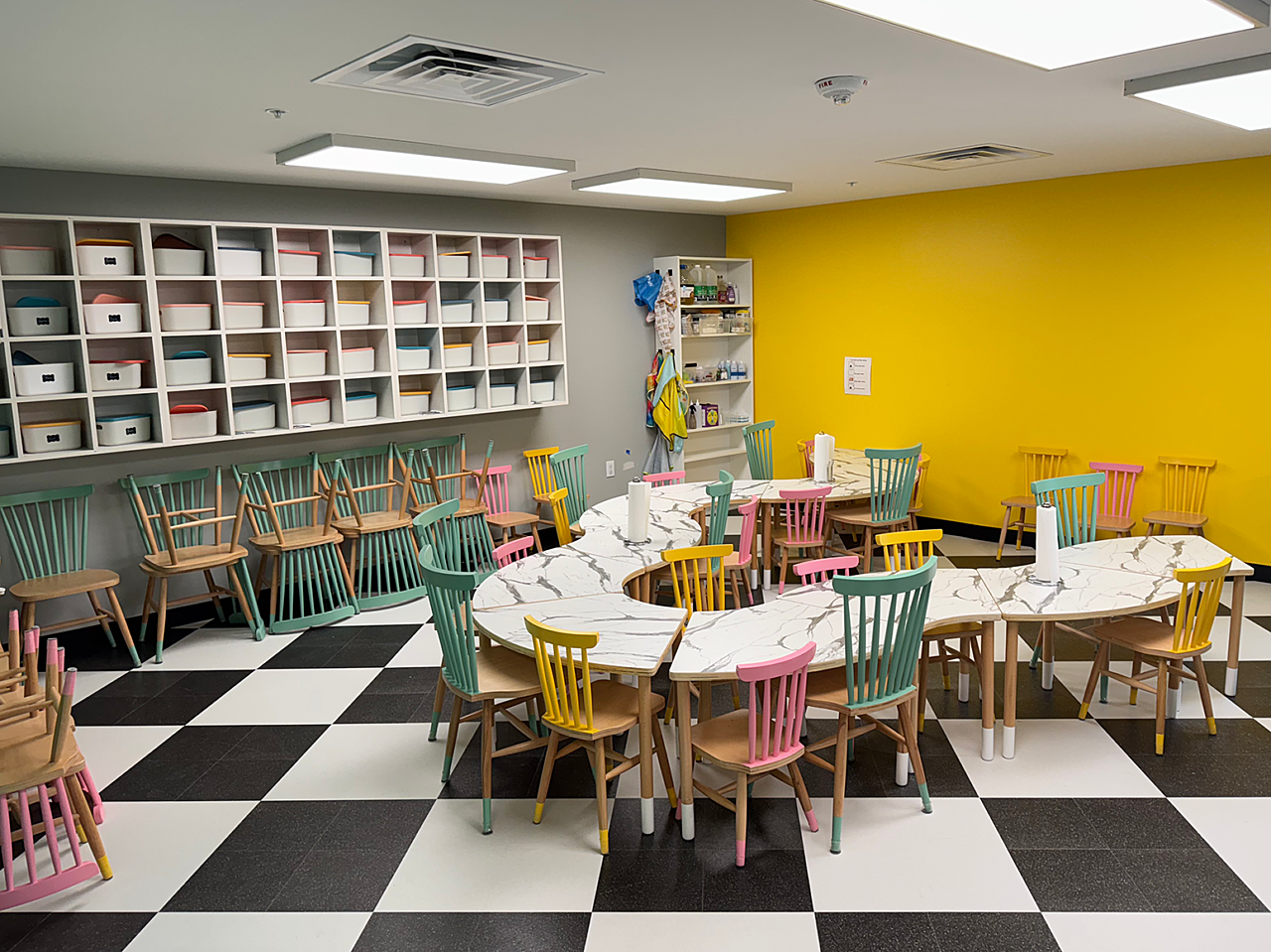 Riley ABA dining area: Child sized curved tables with green, pink and yellow child-sized wooden chairs. cubbies hang on left wall. Black/white tiled floor