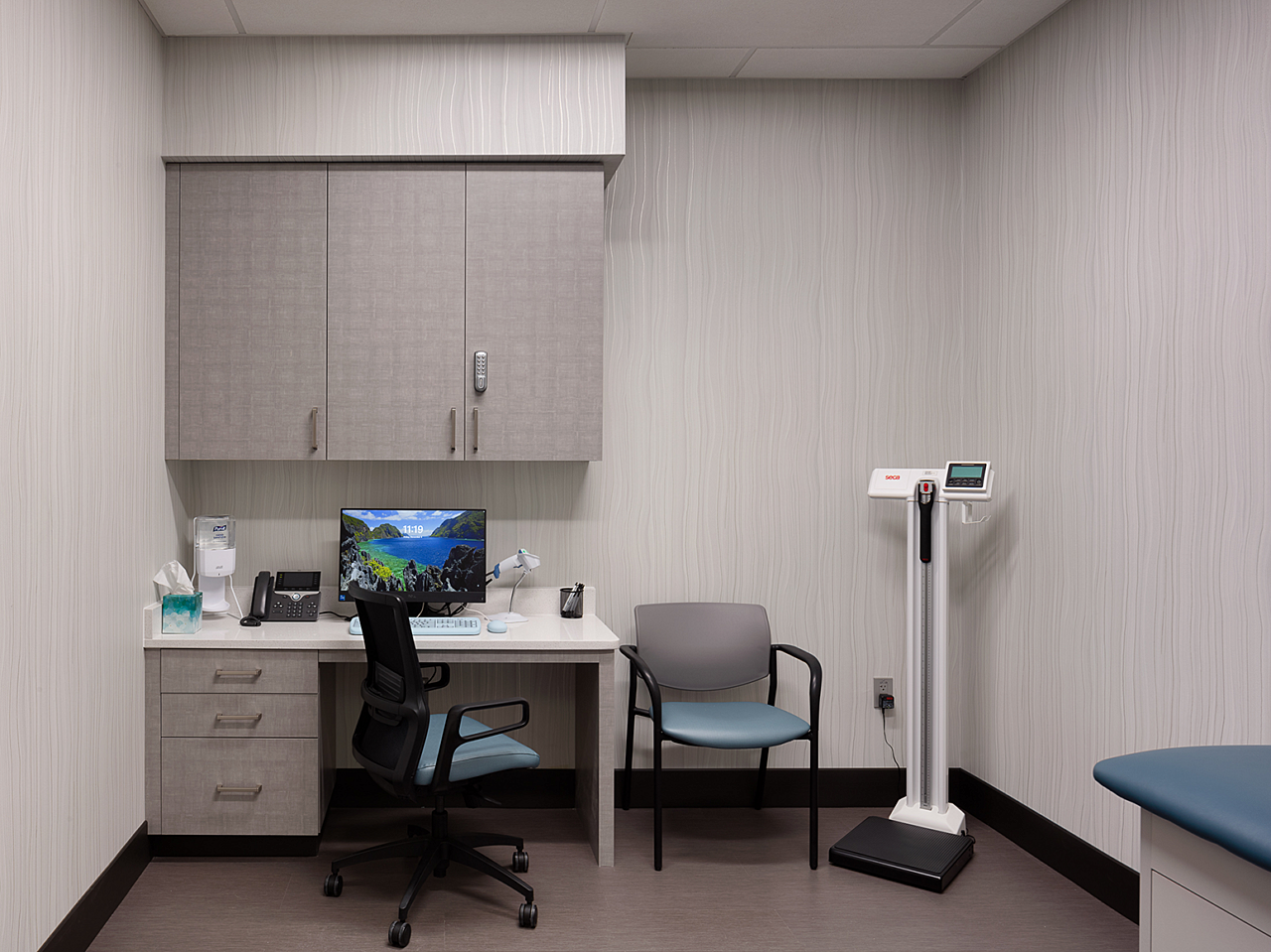 Dr Vince Clinical Research-grey patient exam room with an blue cushioned exam table, side and rolling chair, scale, and desk with laptop and storage.
