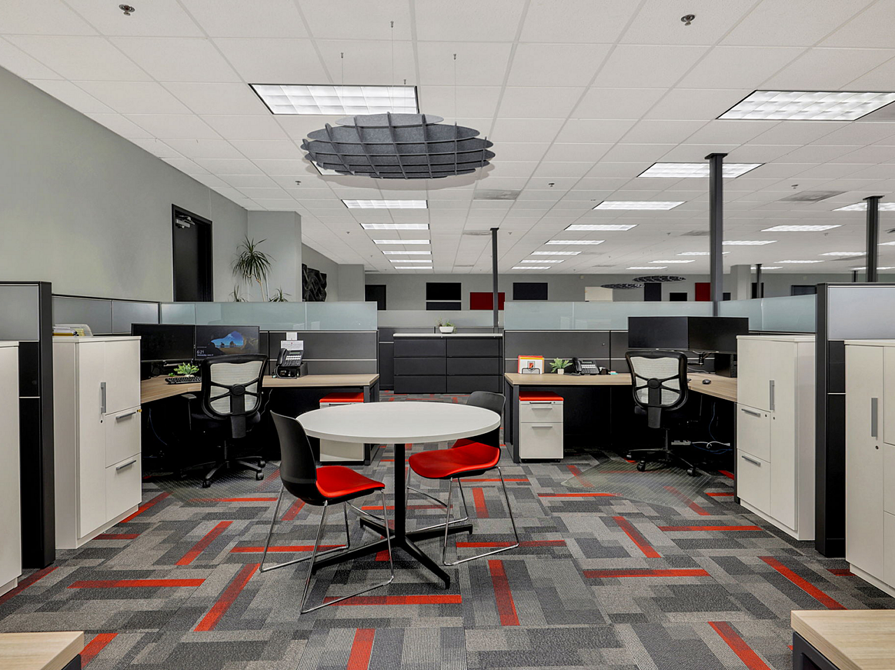 Quad workstation area with small round table and black/red chairs. Storage towers and lateral files. Bold patterned carpet with red and grey rectangles.