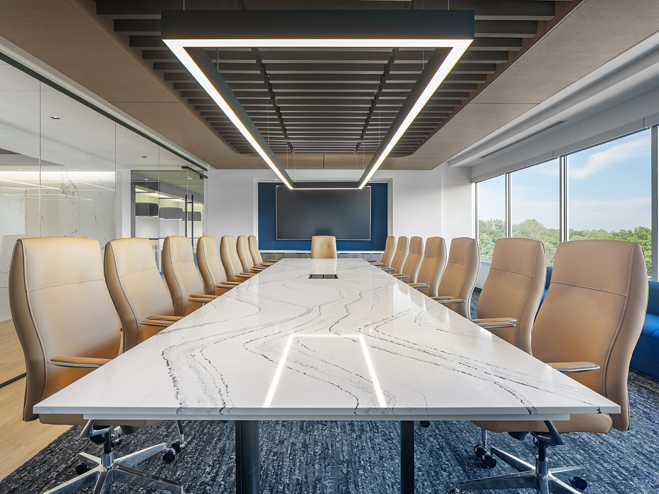 Boardroom with 15 pale gold high-back conference chairs around a white, marble table. Blue bench lines the windows. Dark wood slats on ceiling above table.
