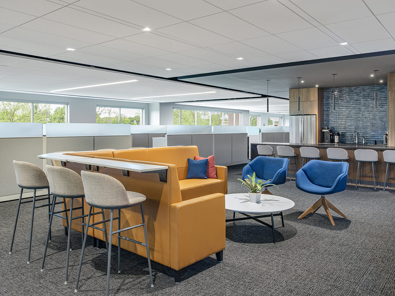 Collab area - L-shaped yellow couch with back rail counter and cream stools. A low white coffee table and two blue 4-prong legged lounge chairs
