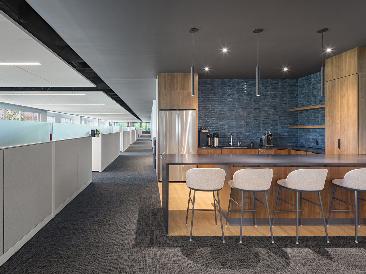 Small kitchenette with a blue tiled backsplash and a marble counter with 4 mid-backed stools across from a corridor-lined with frosted glass cubicle walls
