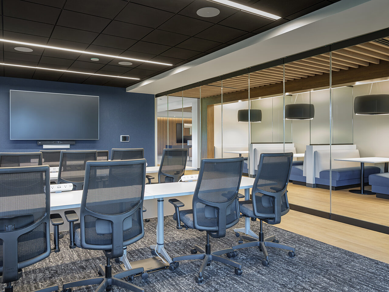 Company training room with long white tables and grey task chairs face a large screen. Outside glass wall is a row of white/blue booths with drop lights
