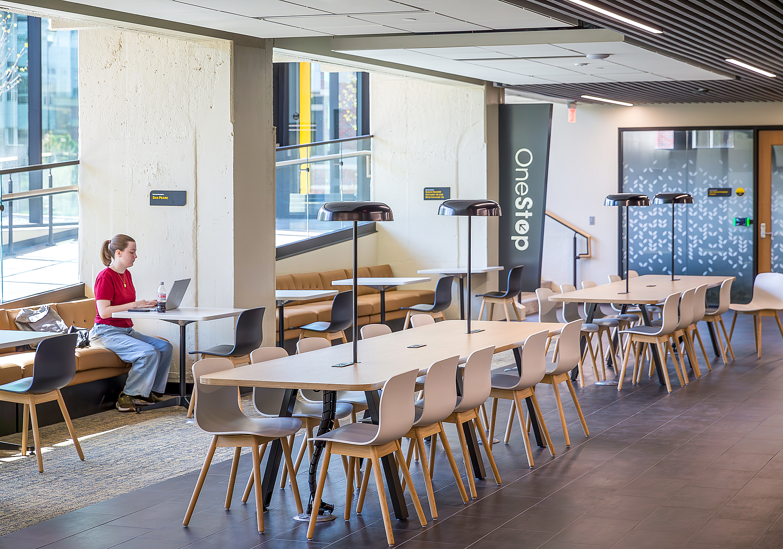 WSU Shocker Center: A female student on a computer in a room w/ banquette seating/cafe tables. Two tables with inserted lamps are in middle of room