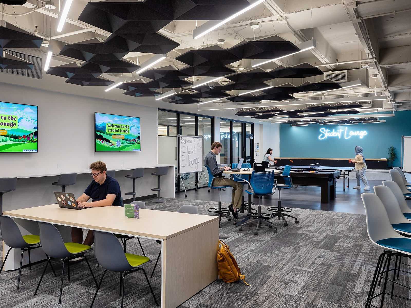 Student Lounge with table, counter and high-top seating areas. Students work on laptops and play ping pong and billiards at the end of the room.