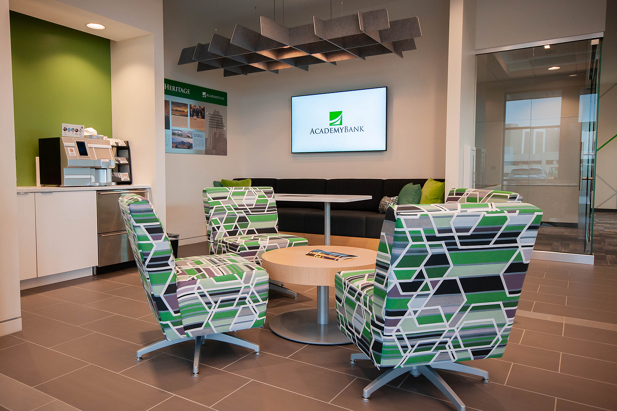 Academy Bank lobby with four green/black/white abstract patterned chairs and coffee table. TV with logo on wall and banquette seating underneath it.