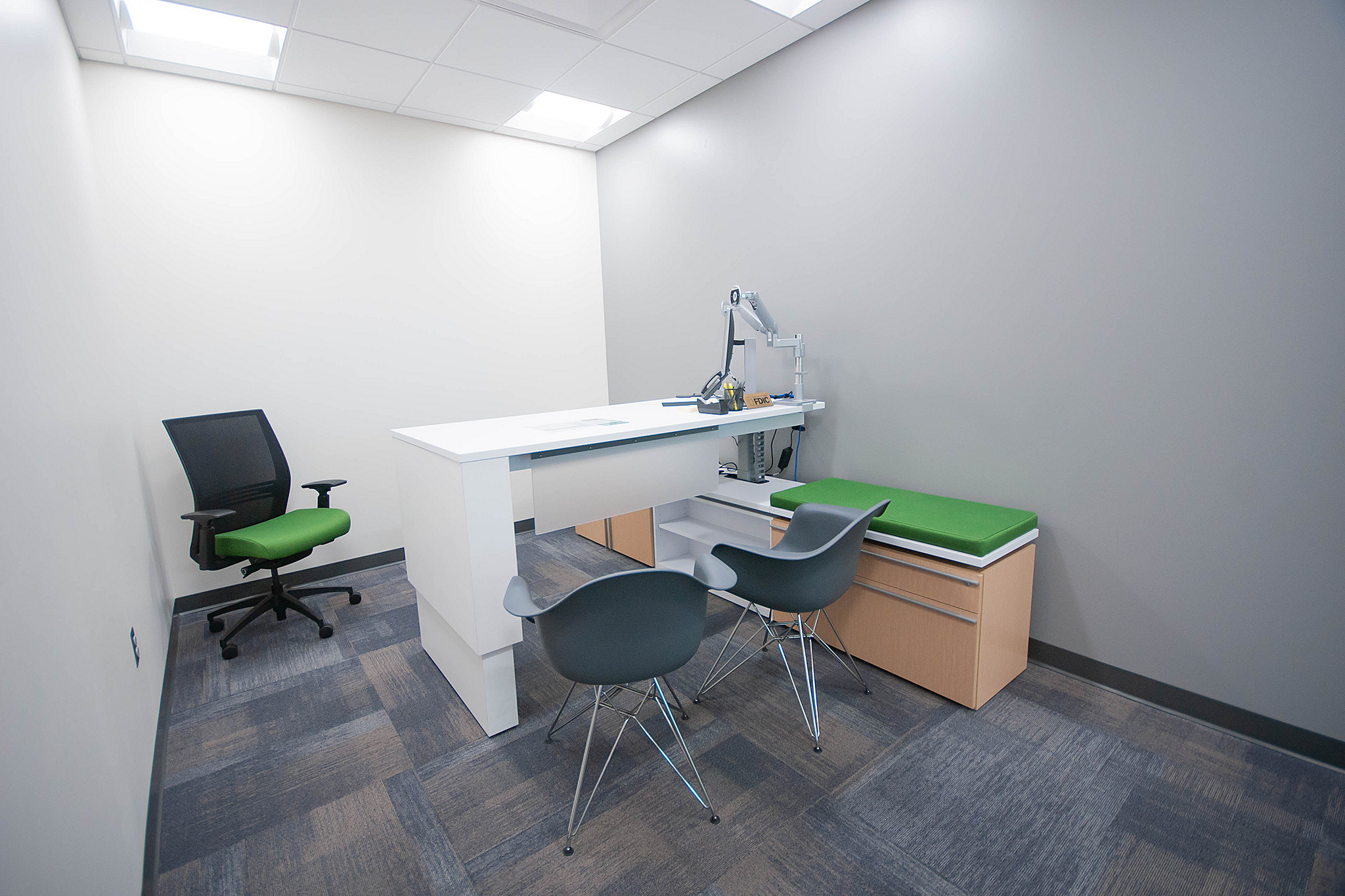 Academy Bank private office with white height-adjustable desk. Black task chair with green seat, two grey guest chairs and wooden storage w/ green cushion
