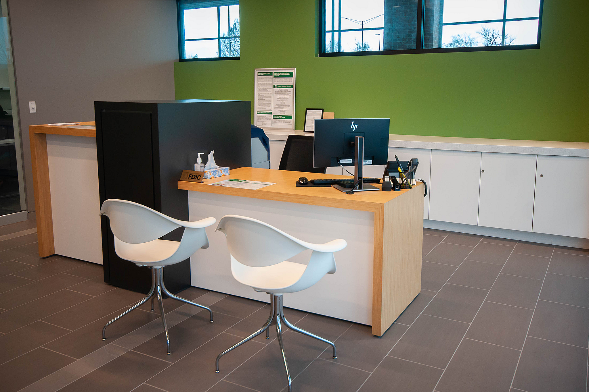 Academy Bank open office cocunter with two white modern guest chairs. Green walls with white cabinets along back wall that has high windows.