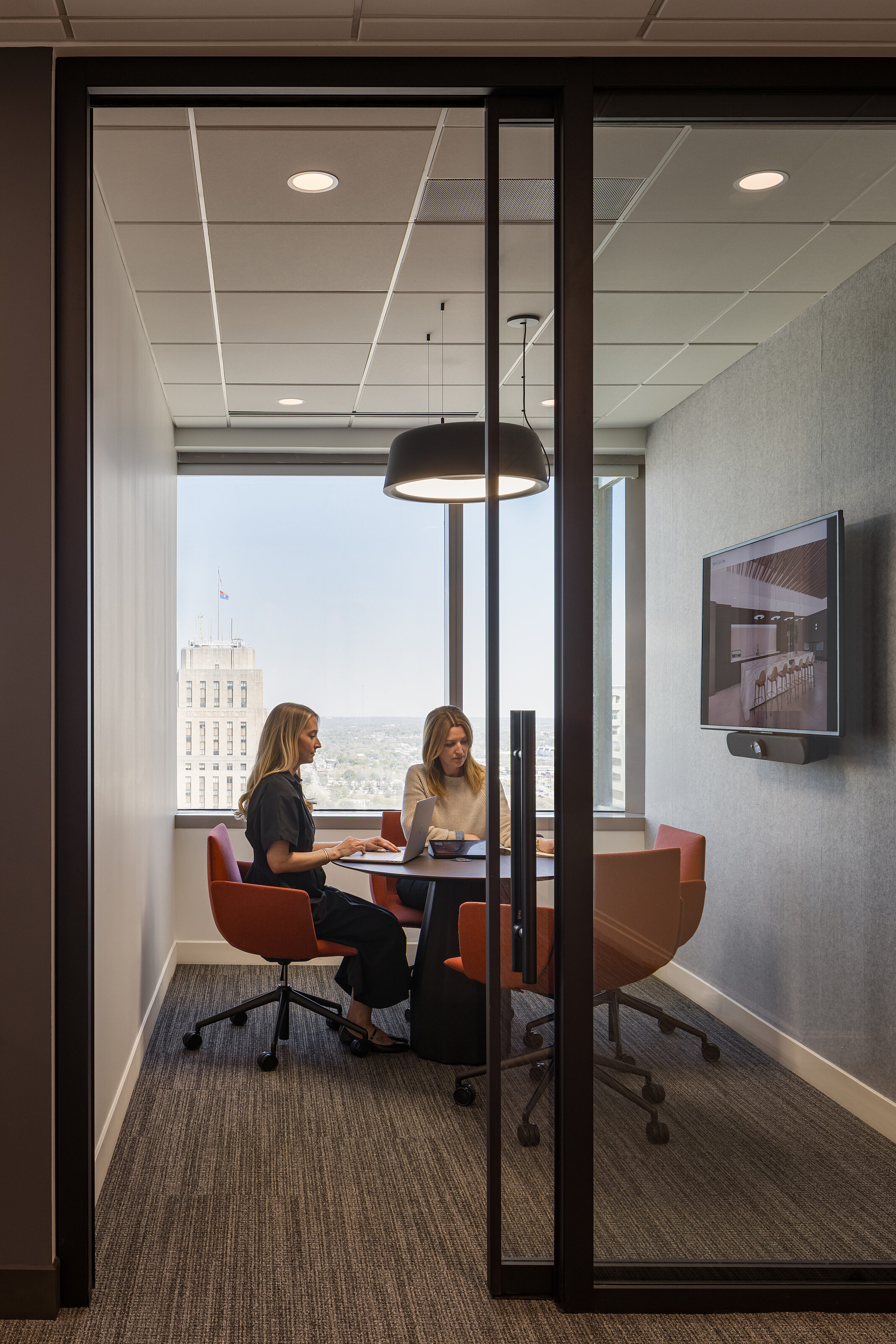 Waypoint: 2 blonde women collaborate in a small, glass-walled conference room with a round table and rounded, dark orange chairs with casters. Skyline view
