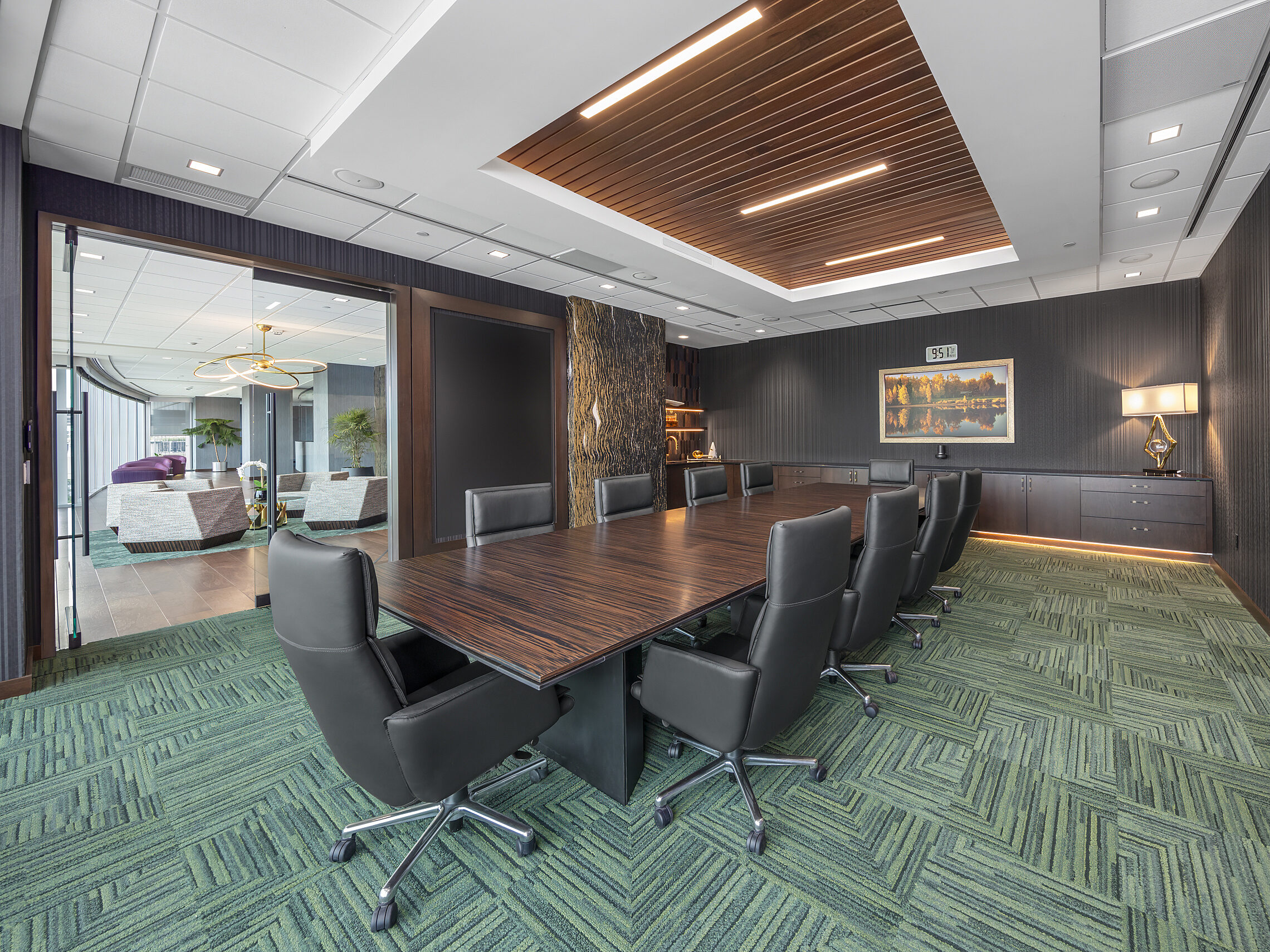 Conference room with textured wood table with high back chocolate chairs, green patterned carpet, dark wood accents and cabinets. Slatted ceiling insert.