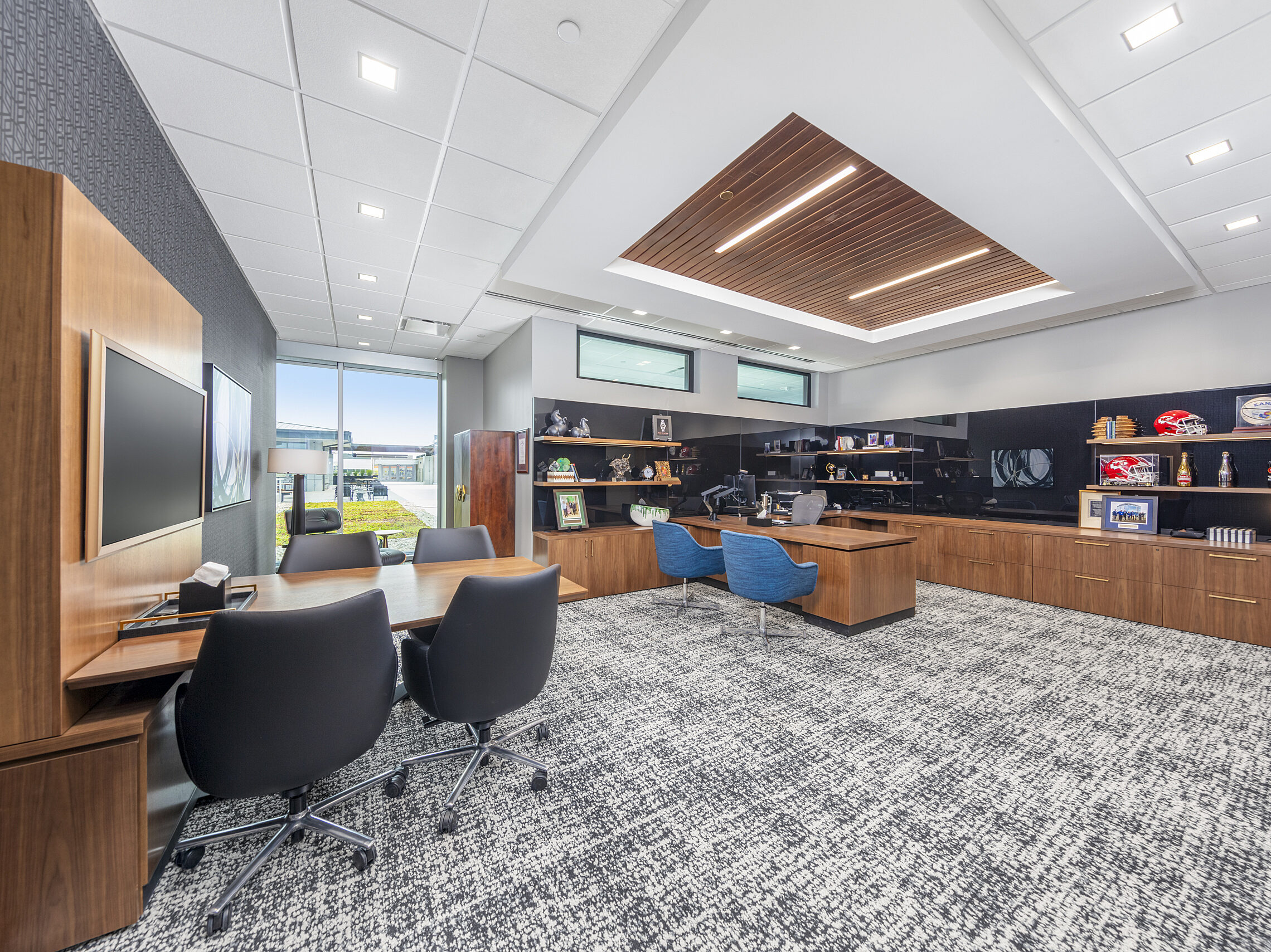 Large executive office: black/white patterned carpet, separate wood desk with blue guest chairs and conference area with black chairs and mounted tv.