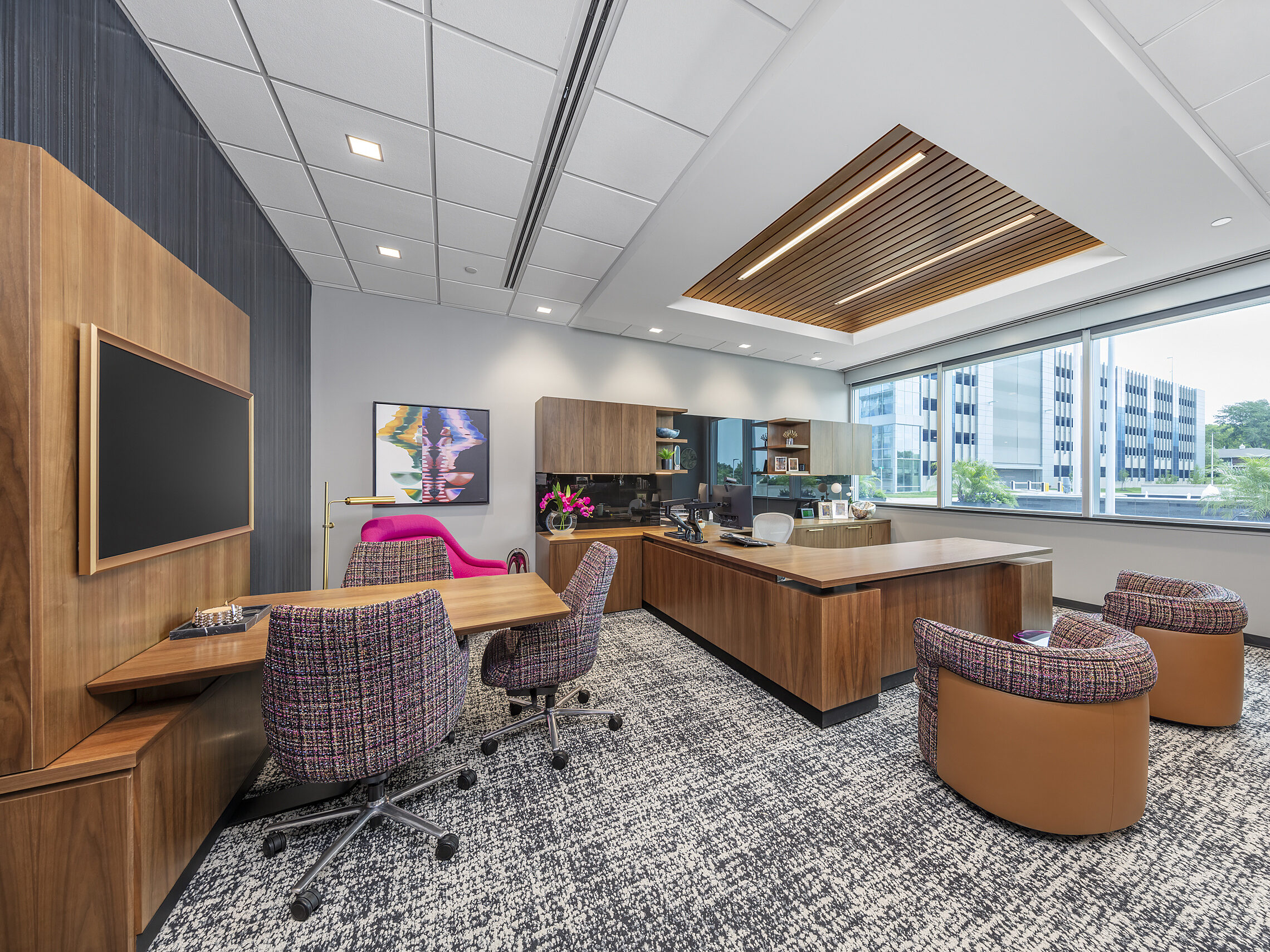 Private office with pink plaid upholstered chairs with leather accents. L-shaped desk unit and separate conference area with affixed table and mounted tv