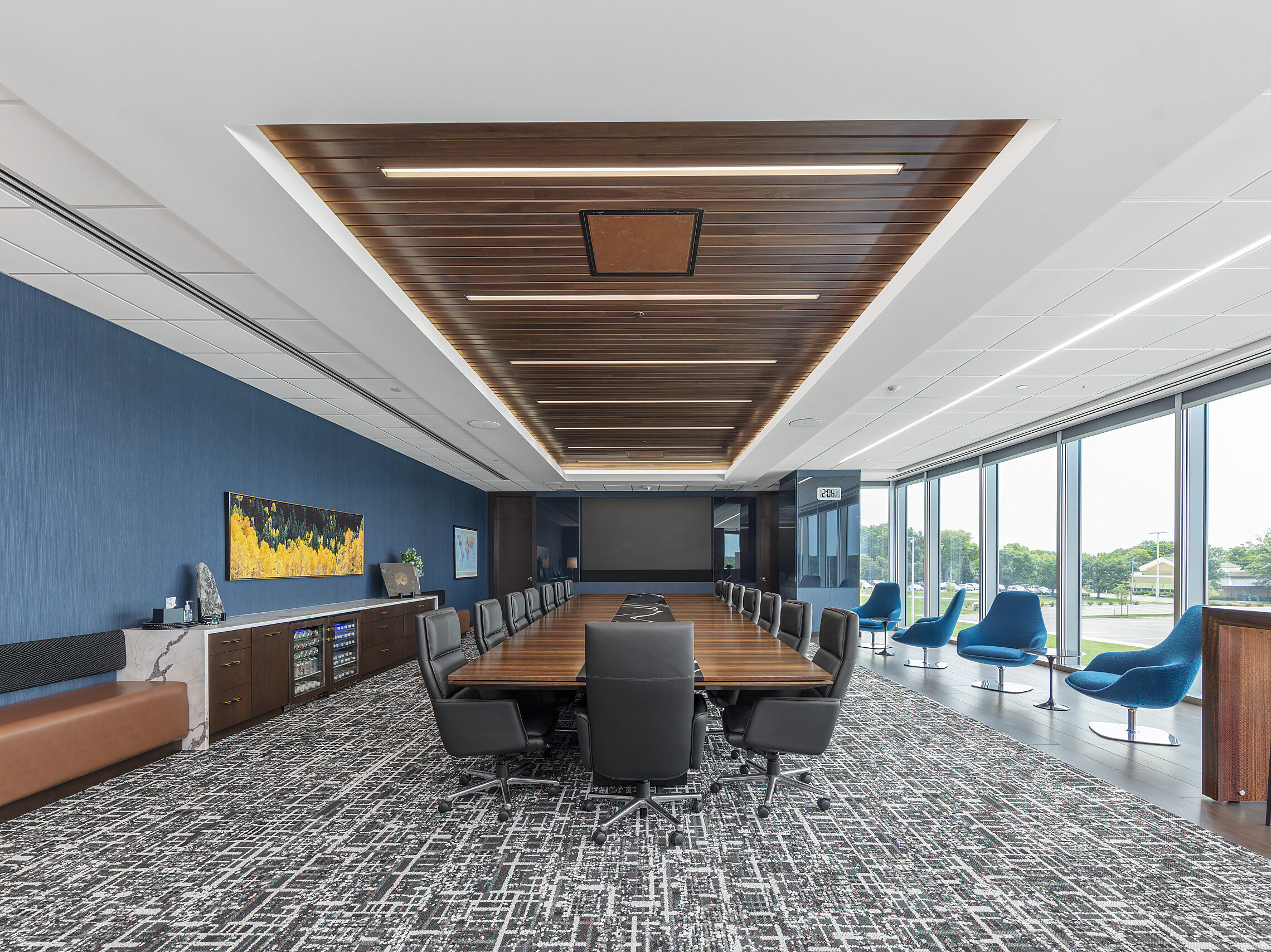 Large conference room table with black marble inset fits 16 people. Black high back chairs. Blue lounge chairs in front of windows. Long cabinet opposite.