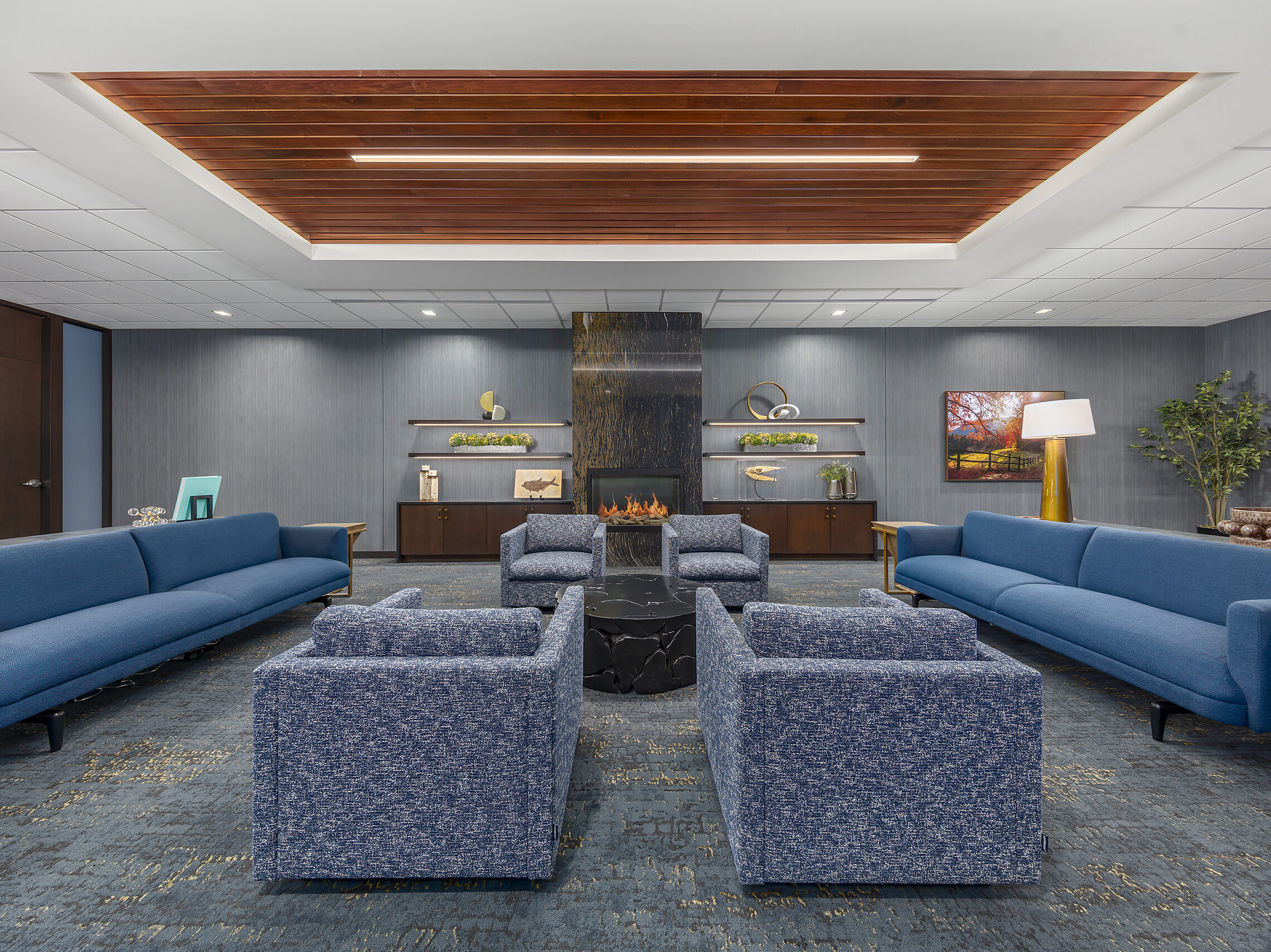 Two long blue sofas and 4 blue patterned armchairs. Narrow textured fireplace with storage cabinets and shelves flanking it. Planked inset ceiling feature