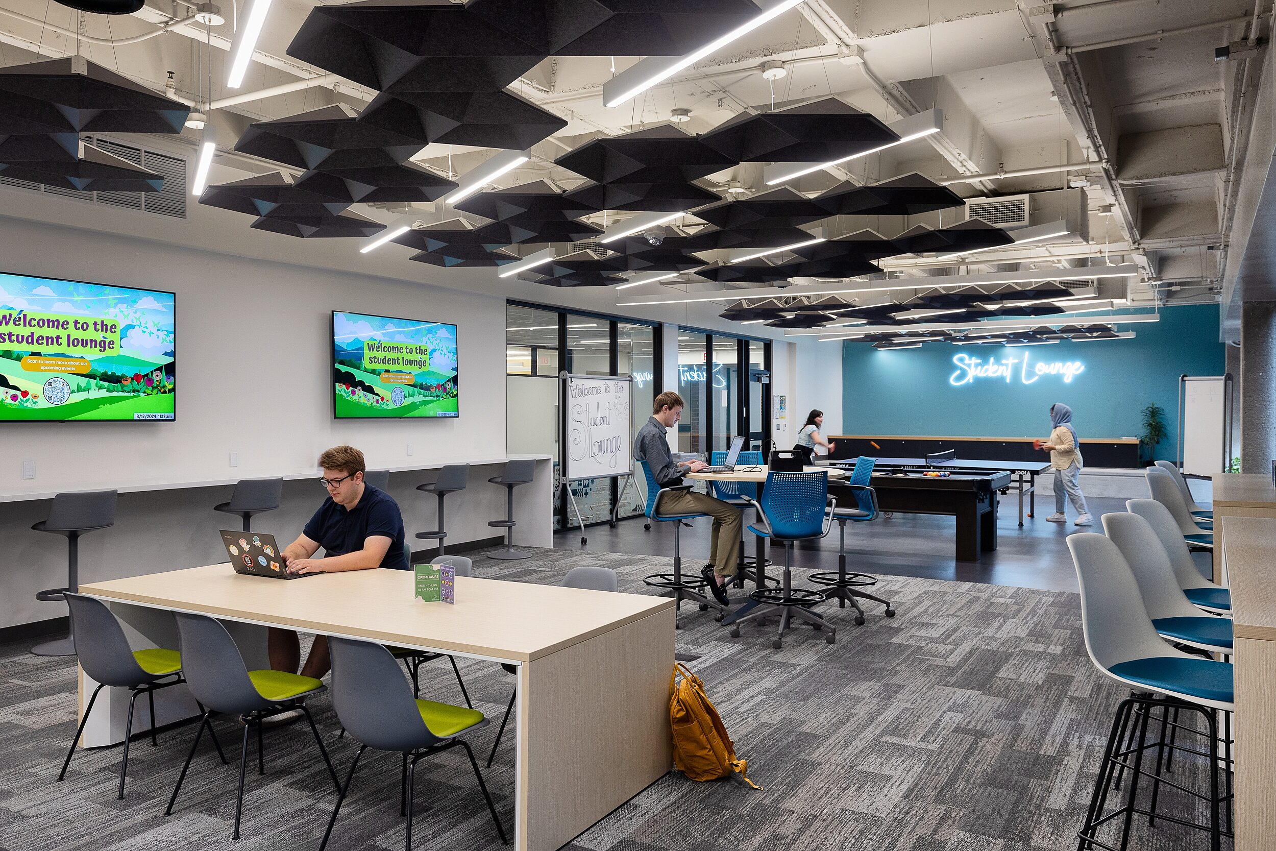 Student Lounge with table, counter and high-top seating areas. Students work on laptops and play ping pong and billiards at the end of the room.