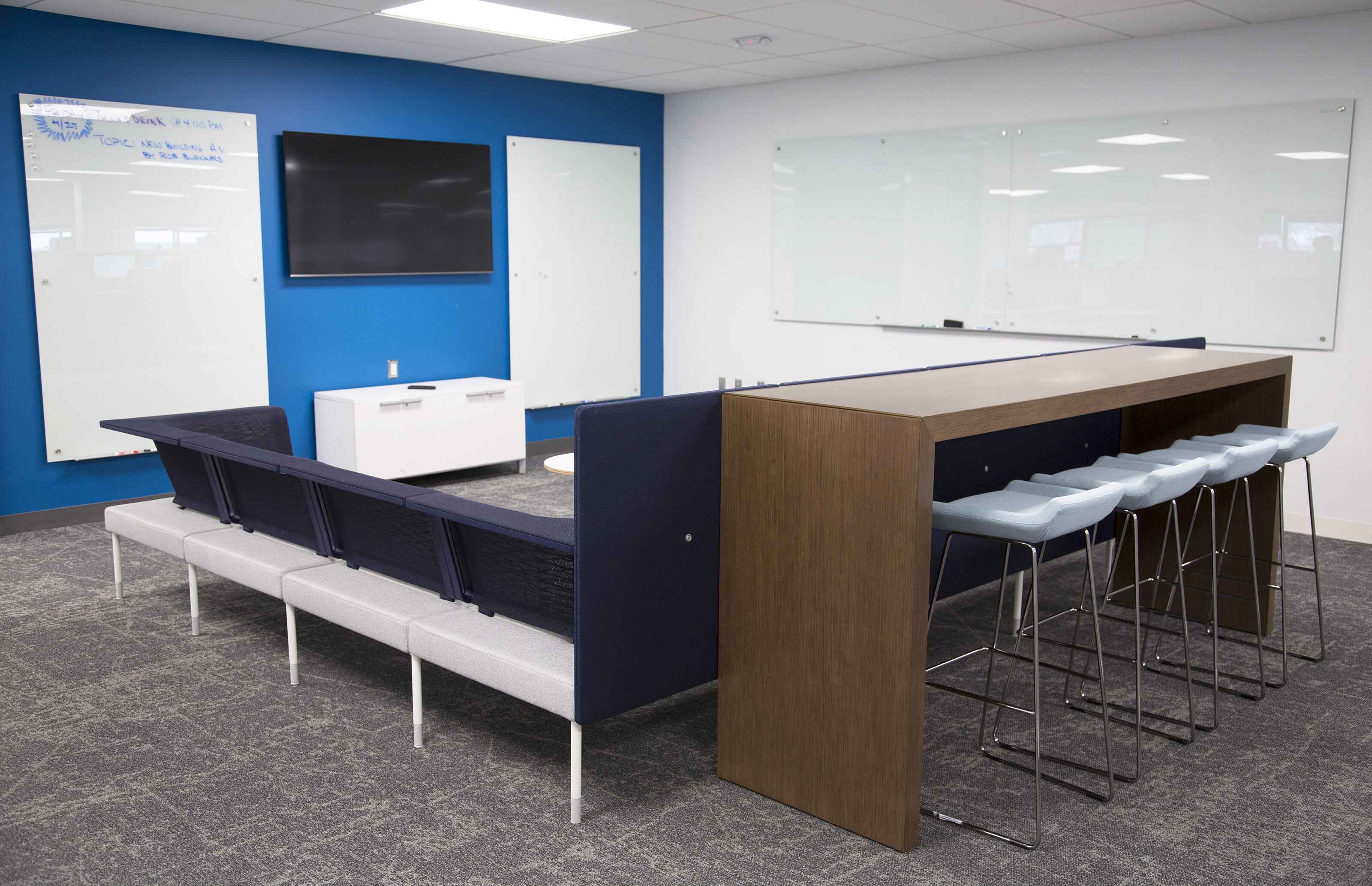 Lockton-collaborative area with u-shaped couch with a wooden waterfall counter and grey stools. 3 white boards and a mounted TV on wall.