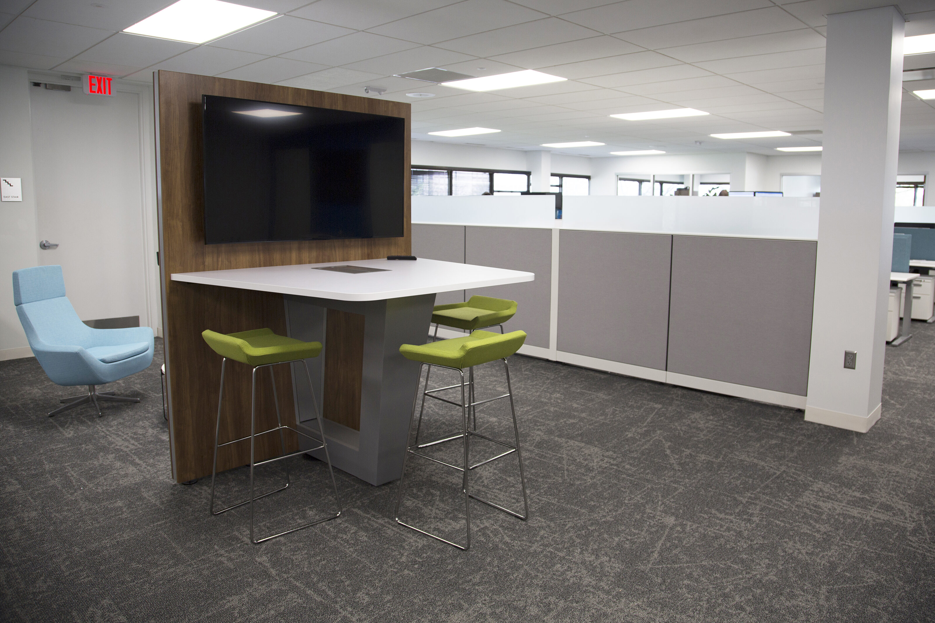 Lockton collaborative area.  Huddle spot with freestanding wall with a mounted TV. Three green stools are around the three sides of the mounted table