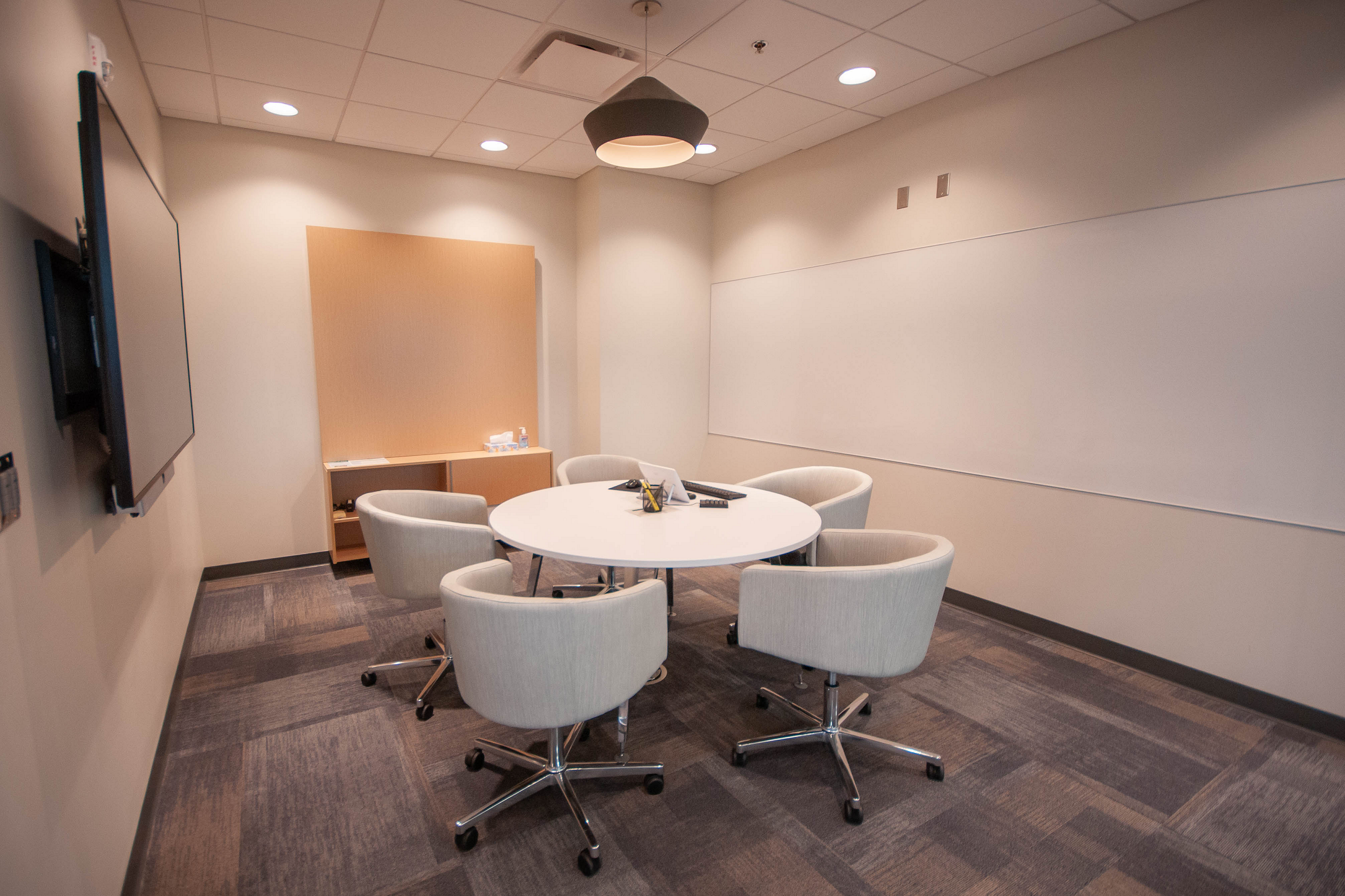 Small Academy Bank conference room. White round table with u-shaped rolling chairs, wall-length white board and tv on two walls. Wooden cabinet on end wall