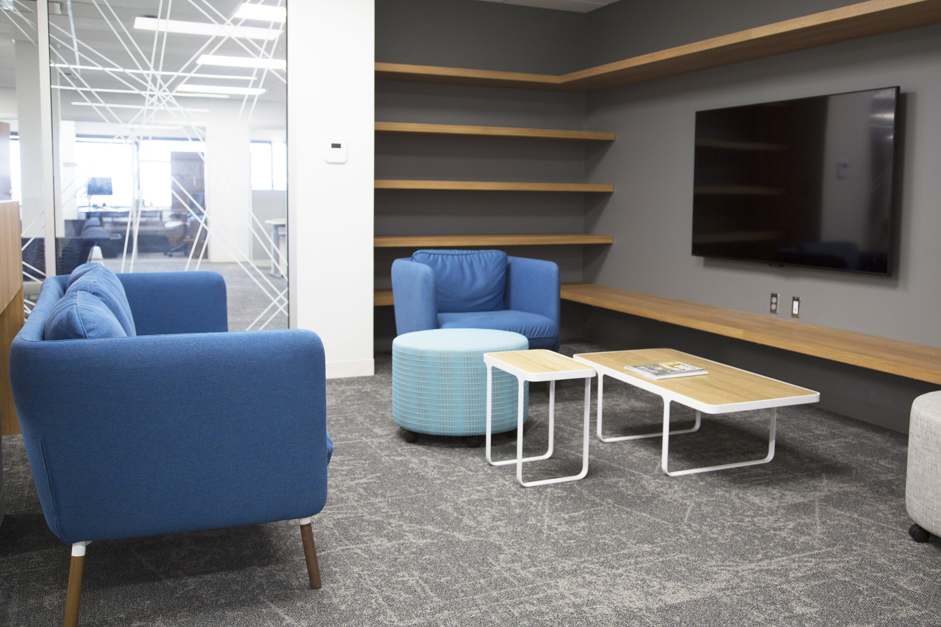 Lockton-small living room with shelving nook and large tv. Blue sofa with legs and lounge chair. White duo coffee tables and blue patterned round ottoman
