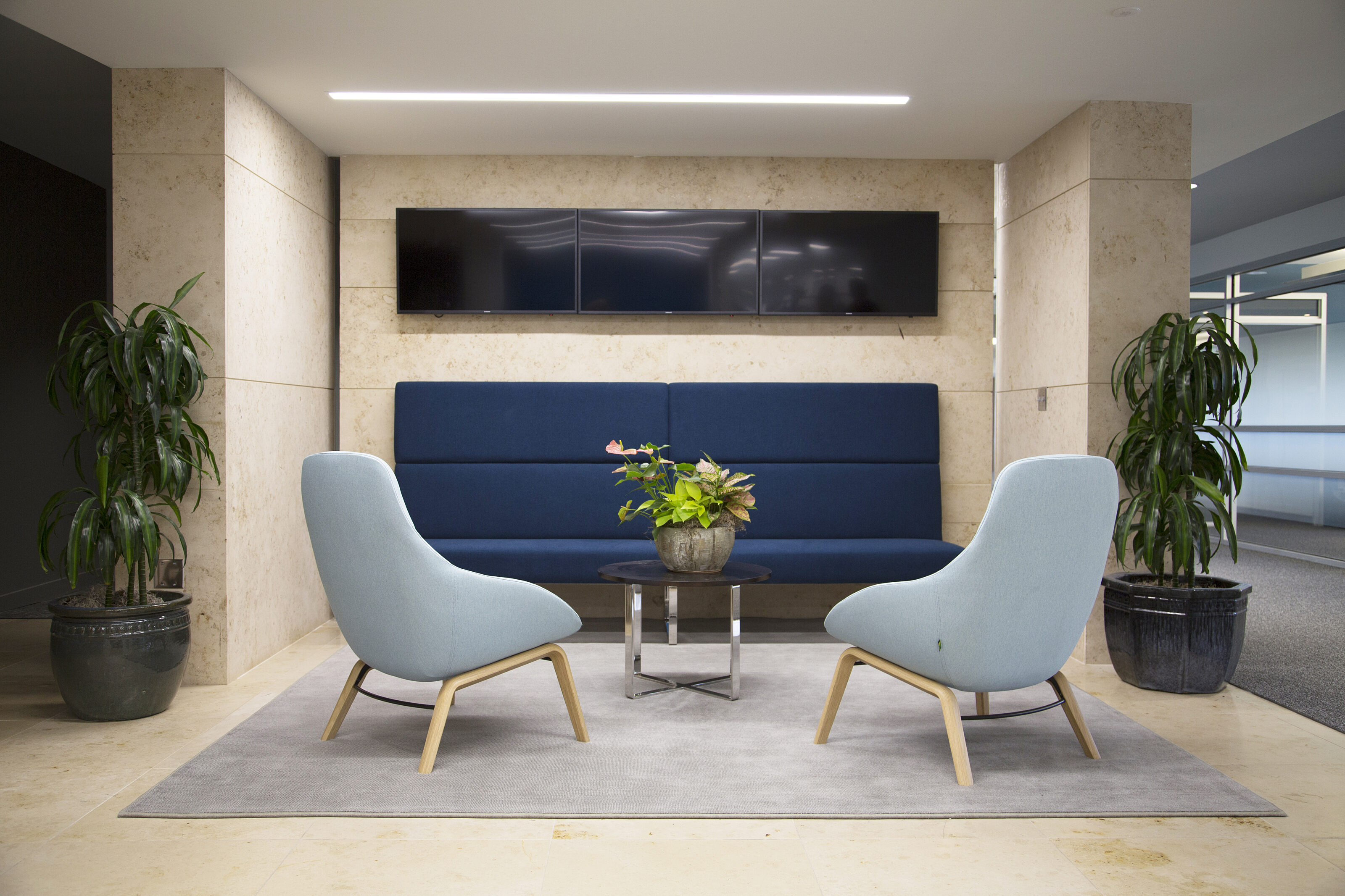 Lockton lounge nook with stone walls and 3 mounted tvs.  Large dark blue banquette sofa and 2 light blue lounge chairs surround small table with plant