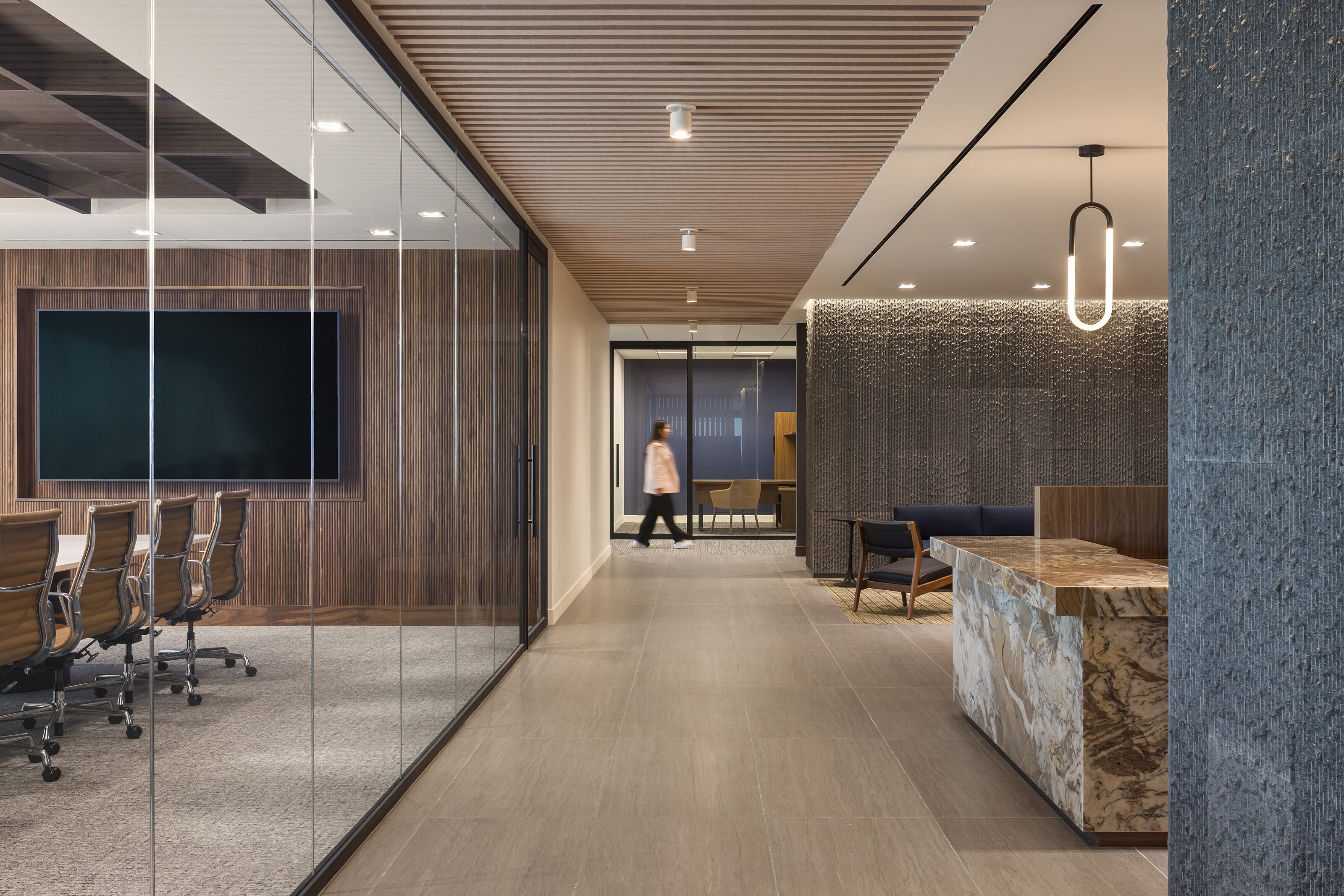 Waypoint: Hallway view that shows glass walled conference room with camel-colored chairs. On right is a marble reception desk and an area with a navy couch
