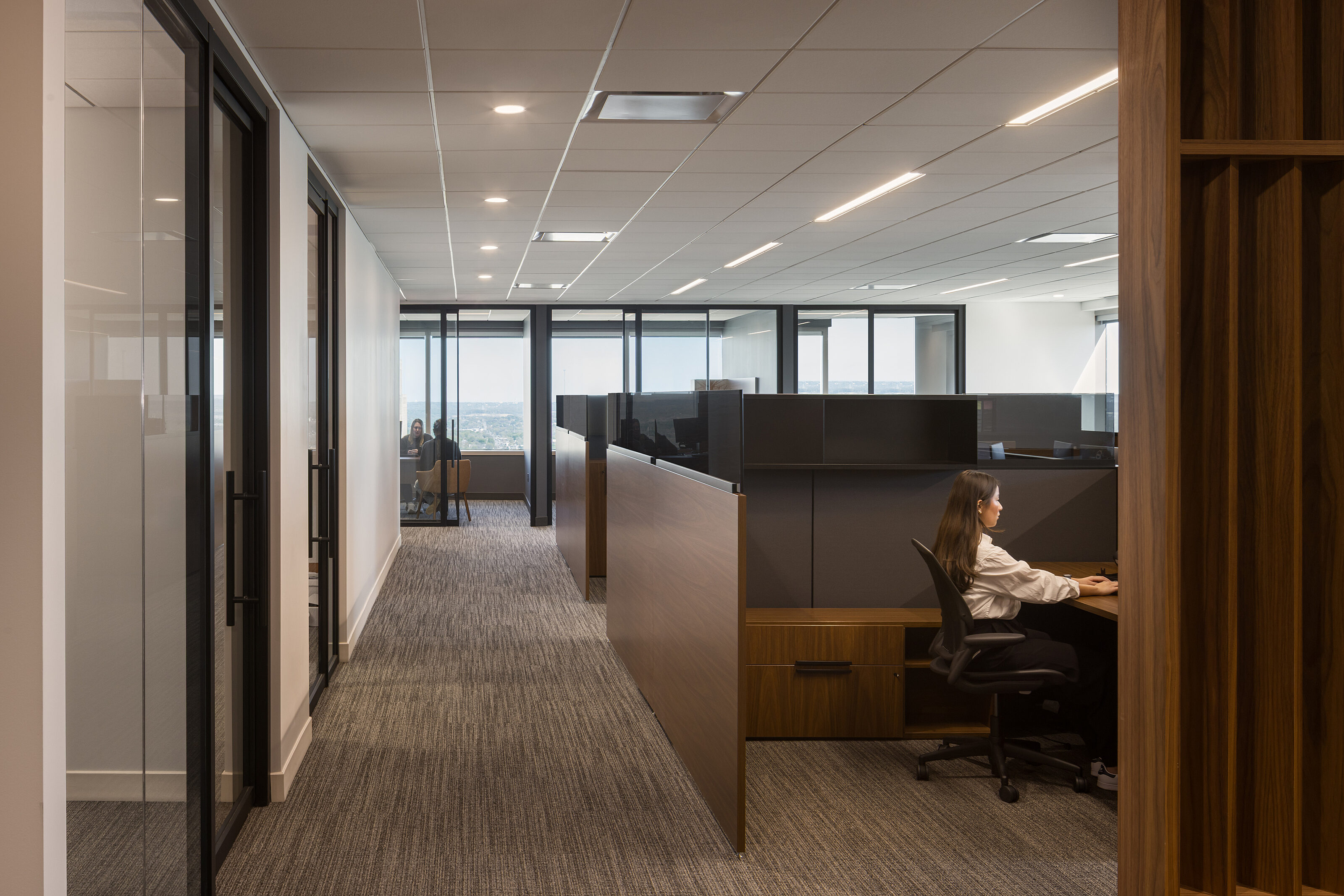 Waypoint: A corridor lined with glass-walled private offices with focus on an area of workstations and a girl at a desk. Dark wood and black accents