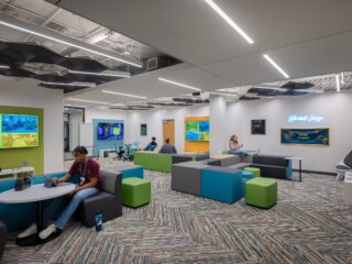 Student Gaming Room, with green, blue and grey tiered cubed soft seating, along with tables and booths. Students play video games and on their phones.