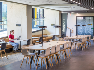 WSU Shocker Center: A female student on a computer in a room w/ banquette seating/cafe tables. Two tables with inserted lamps are in middle of room