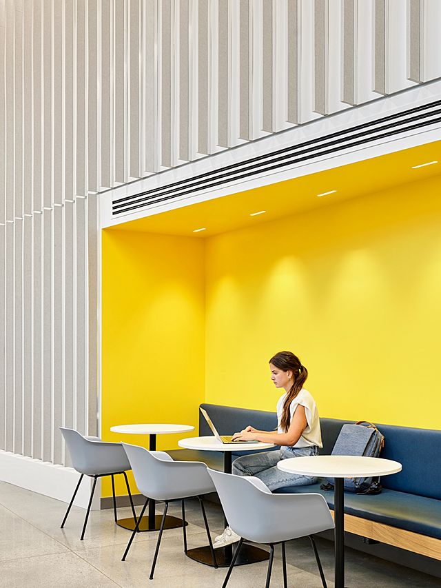 A female college student works on a laptop in a nook area with a vivid yellow paint. She sits on a blue bench with three cafe tables and grey modern chairs
