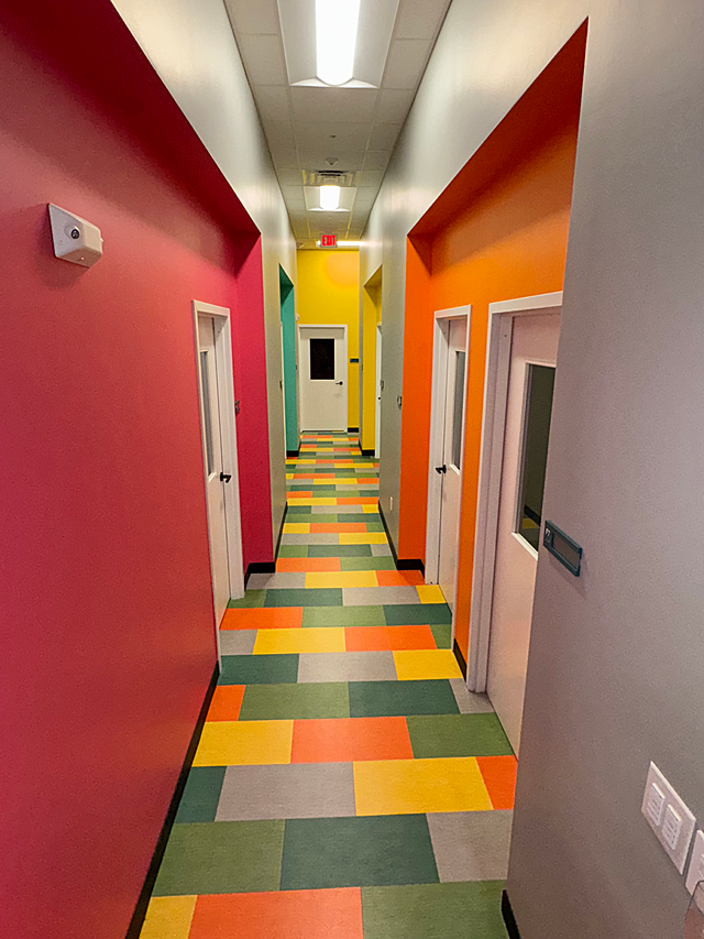 Riley ABA hallway with office doors. Walls painted in solid red, orange, green and yellow down corridor. Carpet tiles in a mix of all the colors