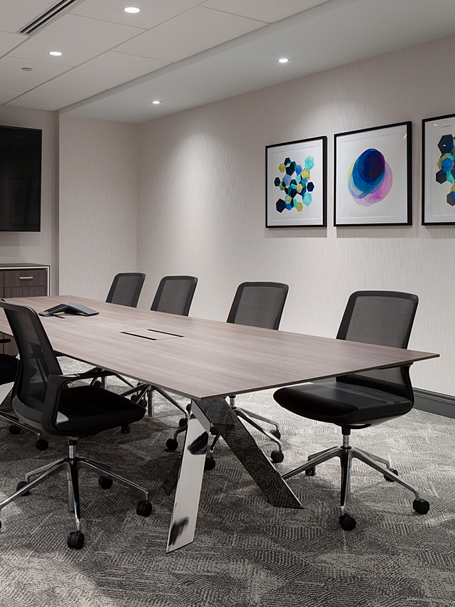 Small conference room with light wood and chrome legs. Surrounded by black and chrome rolling chairs. Three colorful prints in black frames on the wall.