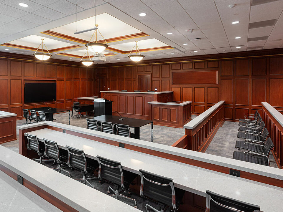Mock courtroom with cherrywood walls. Audience portion contains long white tables and black Eames Aluminum Chairs. Same chairs in jury box to side.