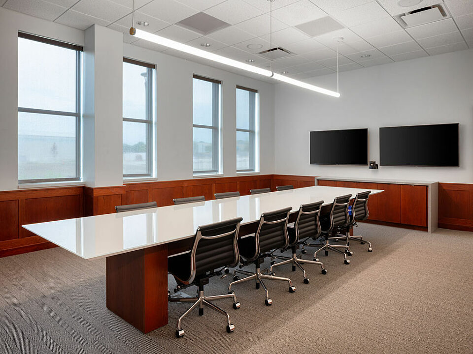 Conference room with cherry wood wainscoting. White surface conference table with black Eames Aluminum chairs. Two mounted TV screens on far end.