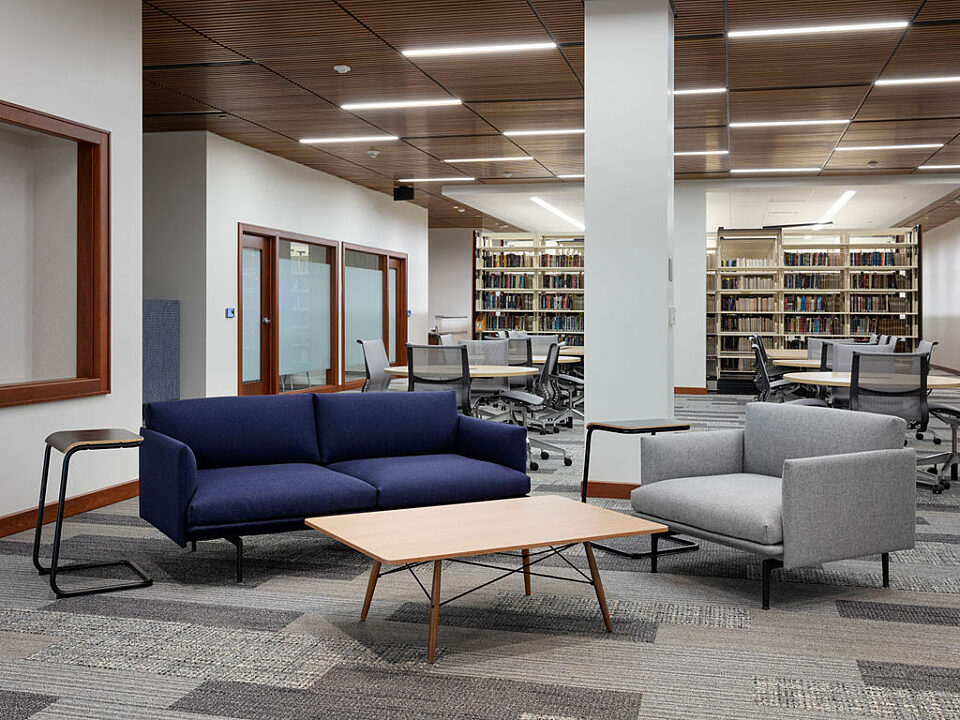 Casual seating area in law library. Blue two-cushioned couch and wide grey lounge chair around wooden coffee table. Two laptop tables help with studying.