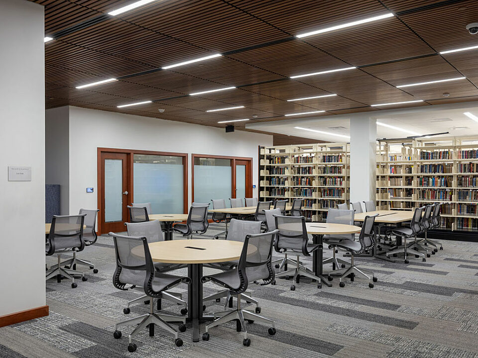 School law library with round and rectangular study tables and rolling high-back task chairs. Multiple shelves filled with books fill the background.