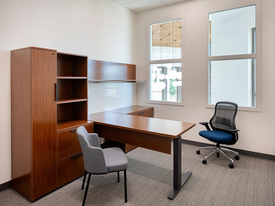 A private office with a wooden, height-adjustable desk with shelving and storage. A blue/black task chair sits near the windows and a grey guest chair