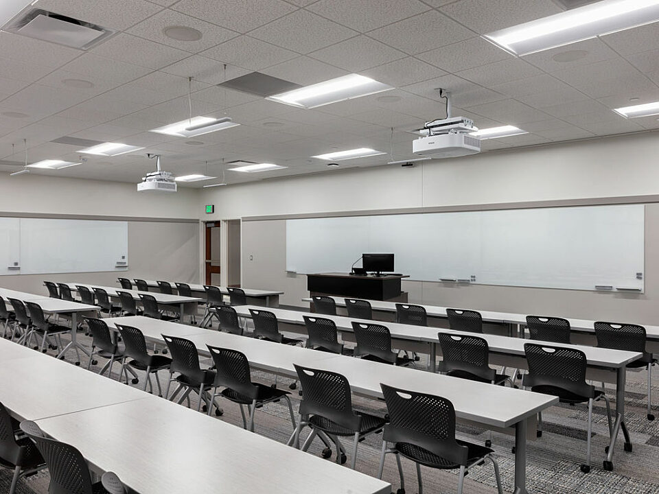 Classroom/training room with two rows of white tables and black chairs. Long whiteboards at front and side of room as well as a black podium