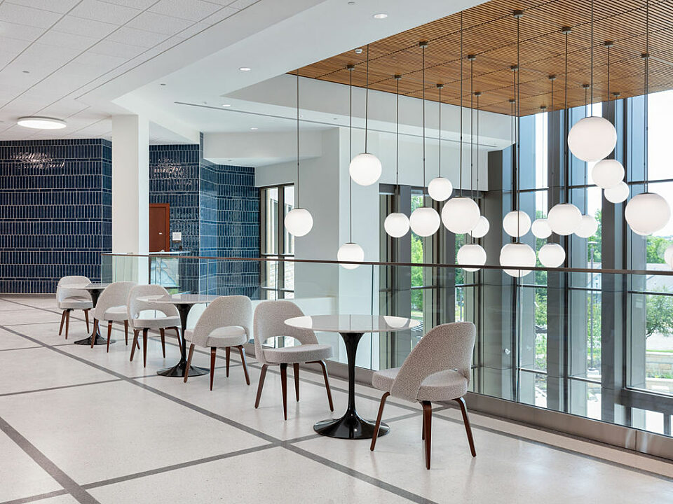 Three sets of white cafe tables with black bases and two grey upholstered chairs sit on a 2nd level balcony. Globe lights hang from wooden ceiling behind