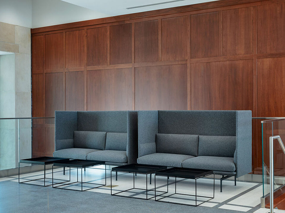 Two grey, high-back upholstered booths with four small black square coffee tables with wire legs in front of a dark wooden accent wall.