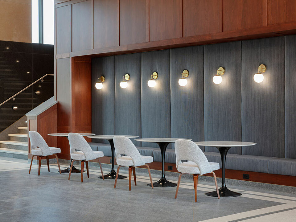 Grey padded banquet nook with seating. Four white topped cafe tables are on the other side. Wall panels in the nook have a mounted hanging globe light.