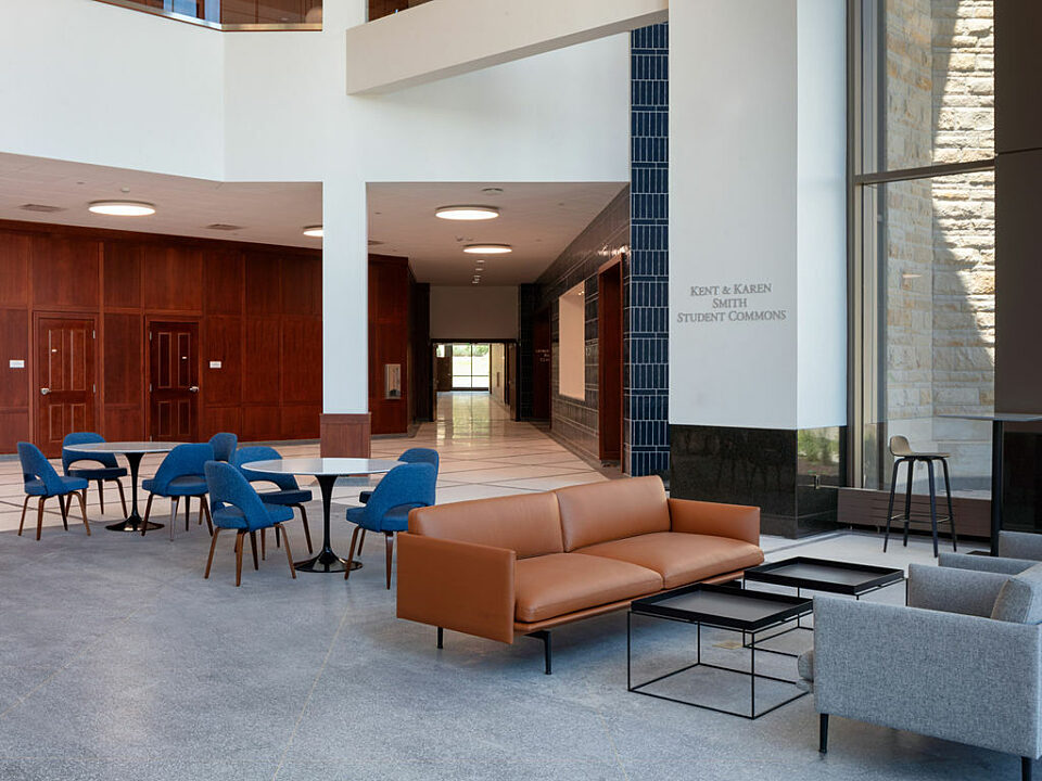 Student commons-hall with brown leather and grey facing sofas with two coffee tables. Behind are round black cafe tables and blue upholstered chairs.